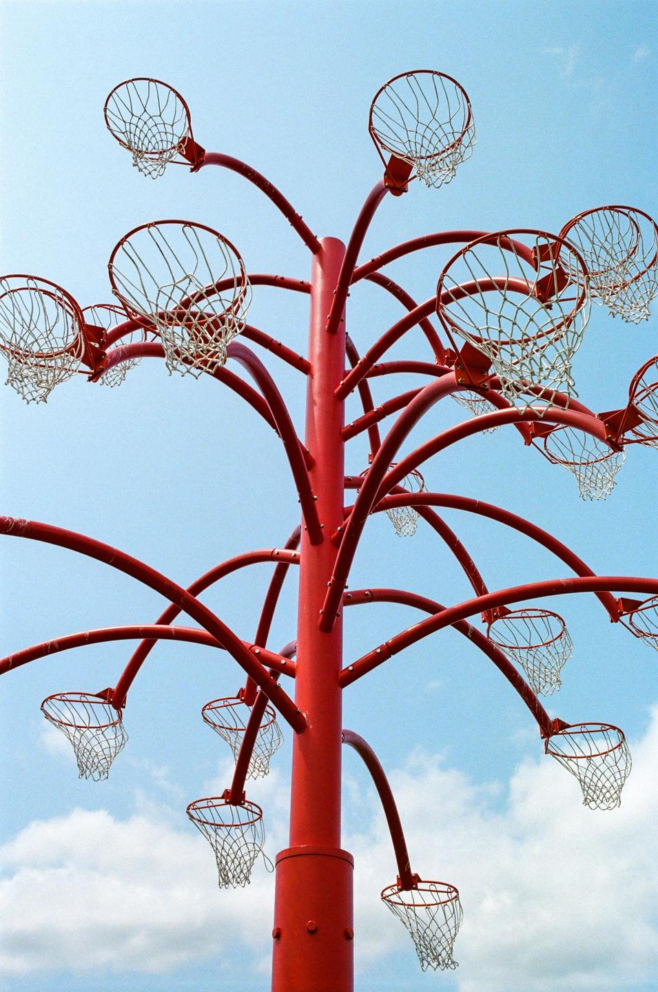 A red pole with multiple basketball hoops extends towards a blue sky with white clouds.