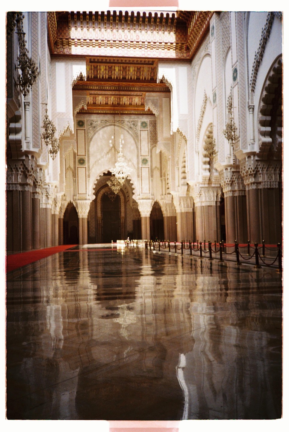A grand hall with ornate arches, chandeliers, and a highly reflective floor leading to a large doorway.