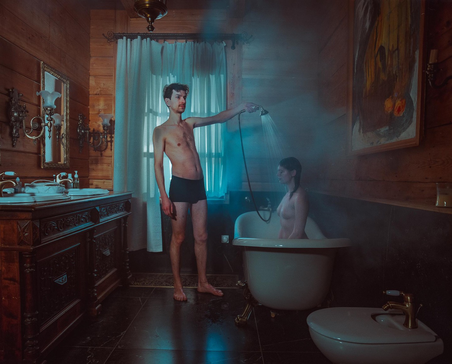 A man showers a woman in a bathtub in a rustic bathroom.
