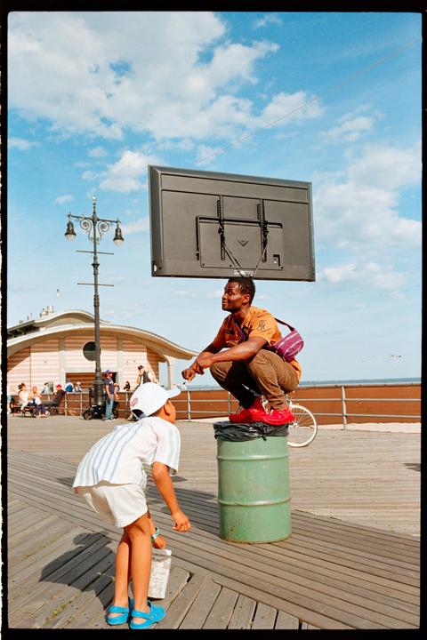 A man squats on a barrel with a television on his head while a boy looks up at him on a boardwalk.