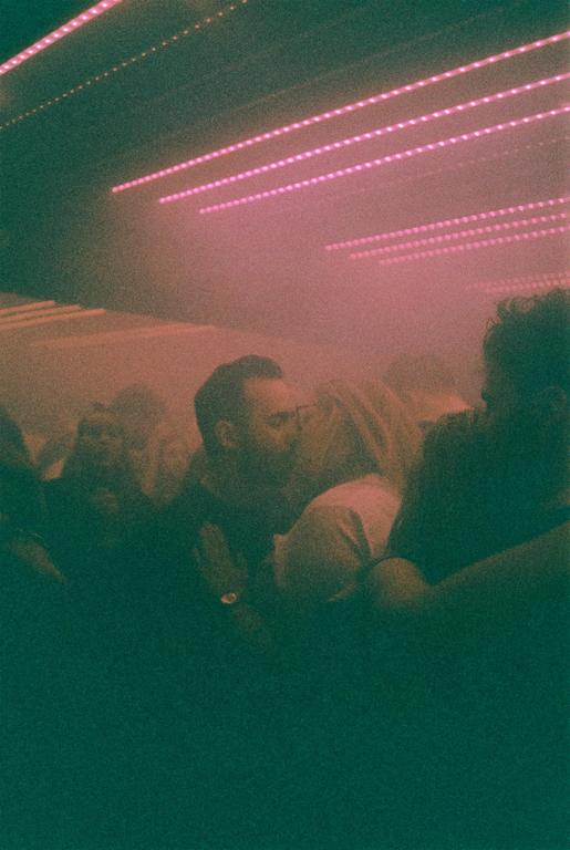 A couple kisses in a dimly lit, smoky room with pink neon lights on the ceiling.