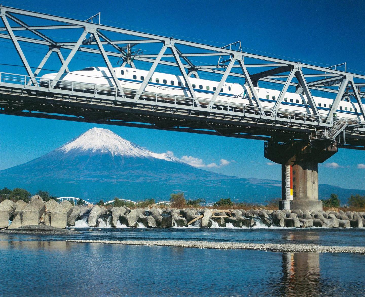 A white bullet train crosses a bridge over a river with a snow-capped mountain in the background.