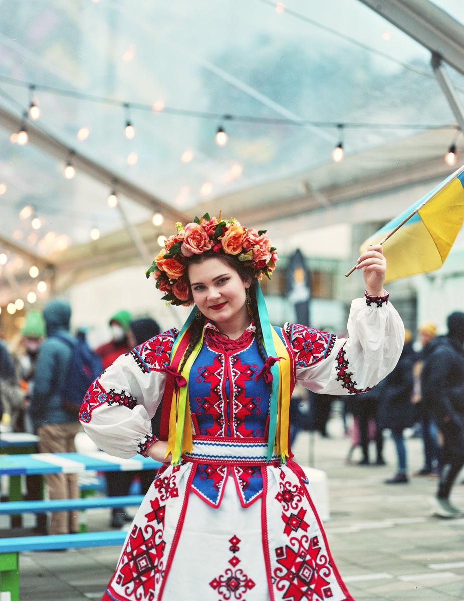 A woman in traditional Ukrainian dress and a floral crown holds a small Ukrainian flag under a tent with string lights.