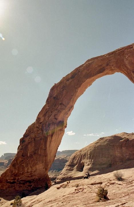 A natural arch of red rock stands tall against a clear sky, with a person sitting on a rock formation below.