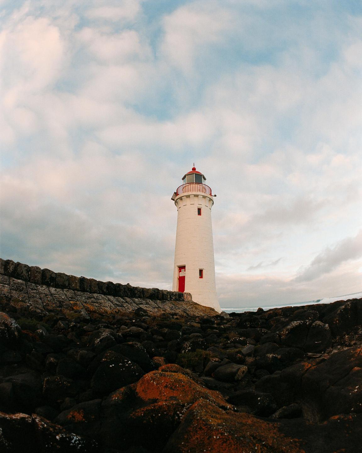 A white lighthouse with a red top and door stands on a rocky shore under a cloudy sky.