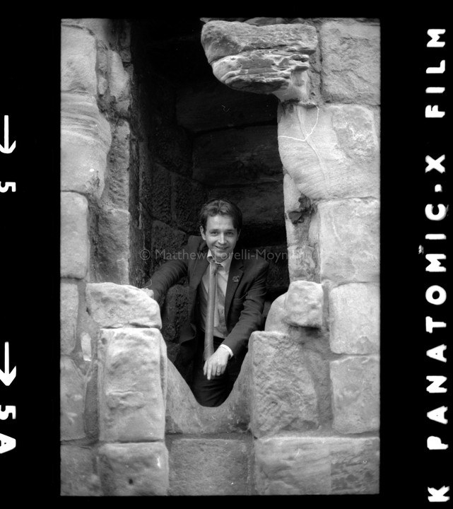 A man in a suit and tie is smiling while looking out from a stone structure.