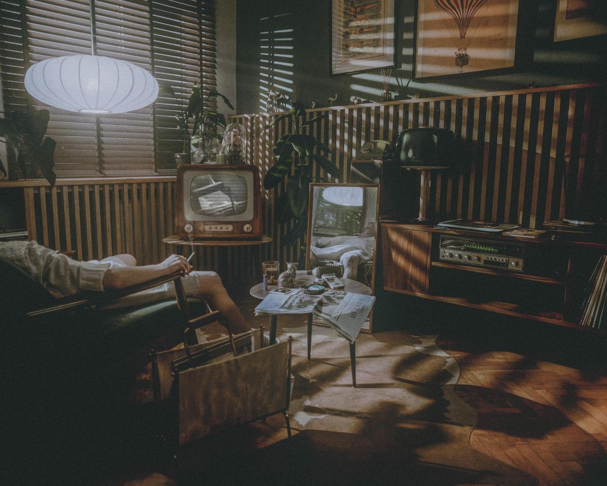 A person reclines in a chair in a living room with a vintage television and stereo system, illuminated by sunlight streaming through blinds.