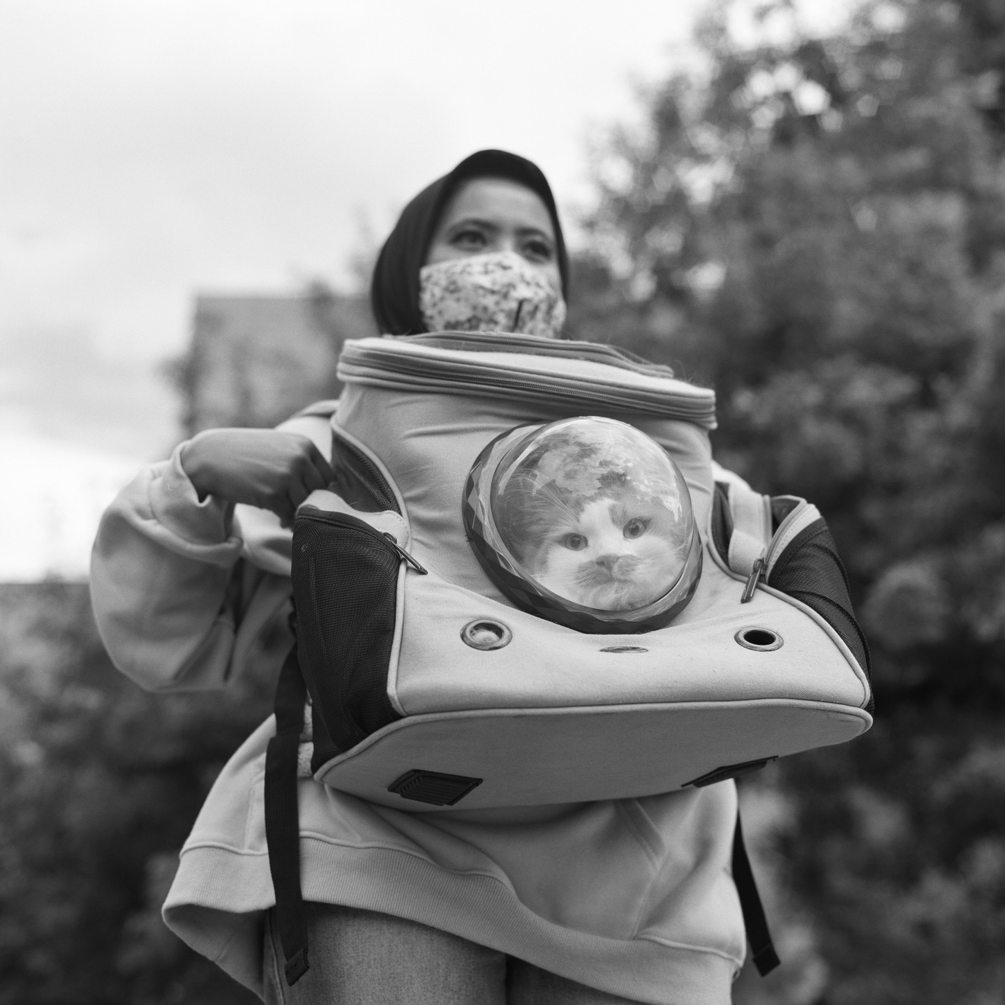 A woman wearing a face mask carries a cat in a backpack with a clear bubble window.