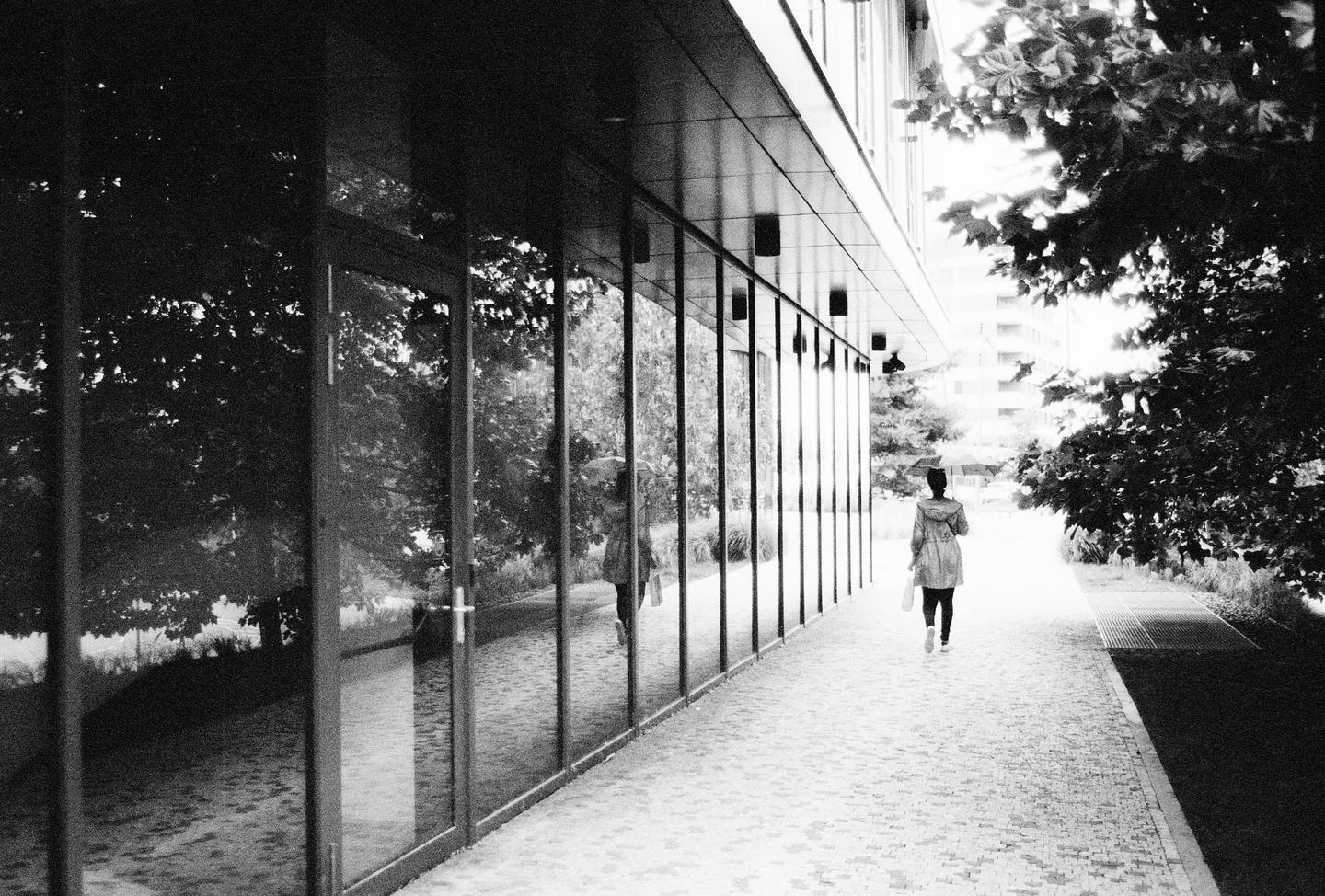 A woman with an umbrella walks along a modern building with reflective windows.
