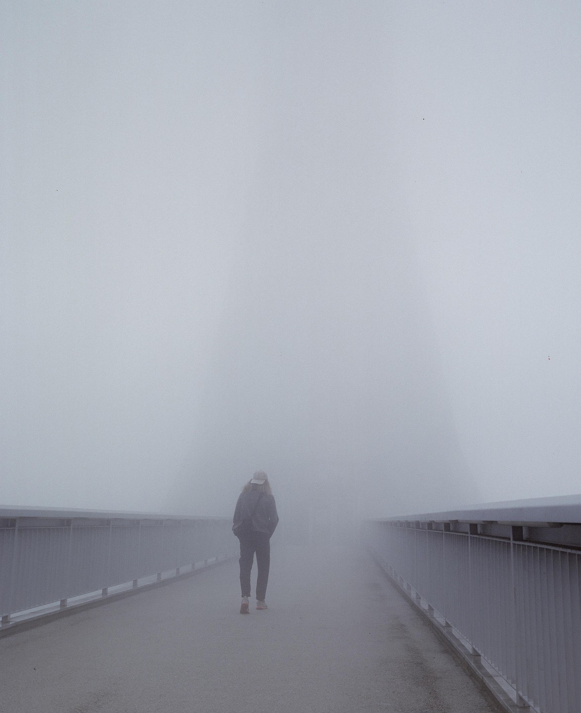 A person walks away from the viewer on a bridge in thick fog.