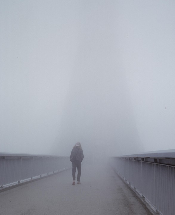 A person walks away from the viewer on a bridge in thick fog.