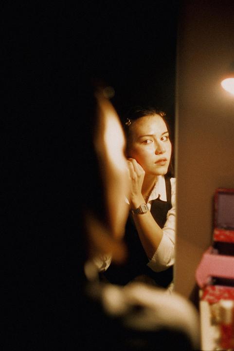 A woman wearing a watch and a vest looks into a mirror while adjusting her earring.