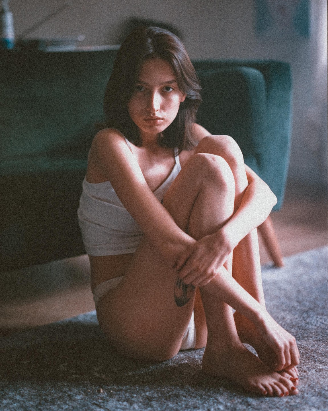 A woman sits on a carpeted floor with her knees pulled to her chest, looking directly at the viewer.