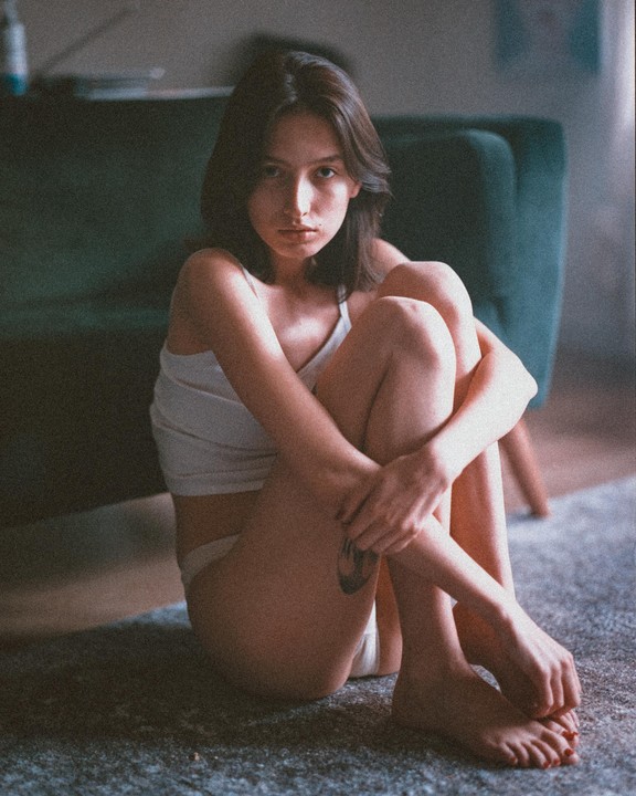 A woman sits on a carpeted floor with her knees pulled to her chest, looking directly at the viewer.