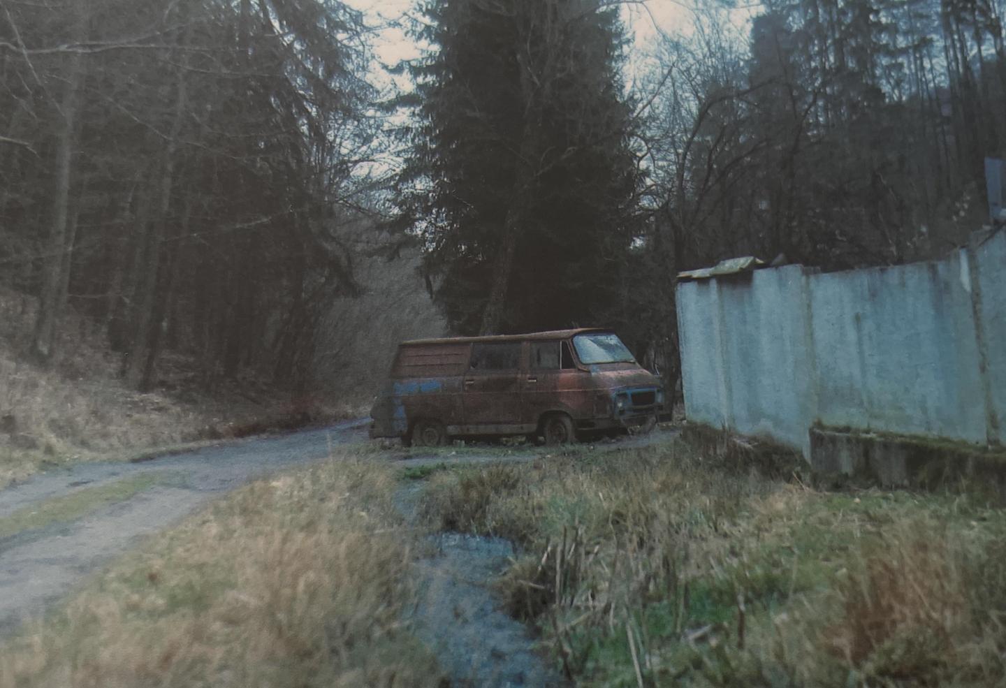 A rusty van is parked on a dirt road next to a concrete wall and a forest.