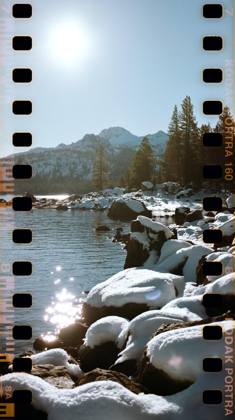 A bright sun shines over a snowy mountain lake with pine trees and snow-covered rocks along the shore.