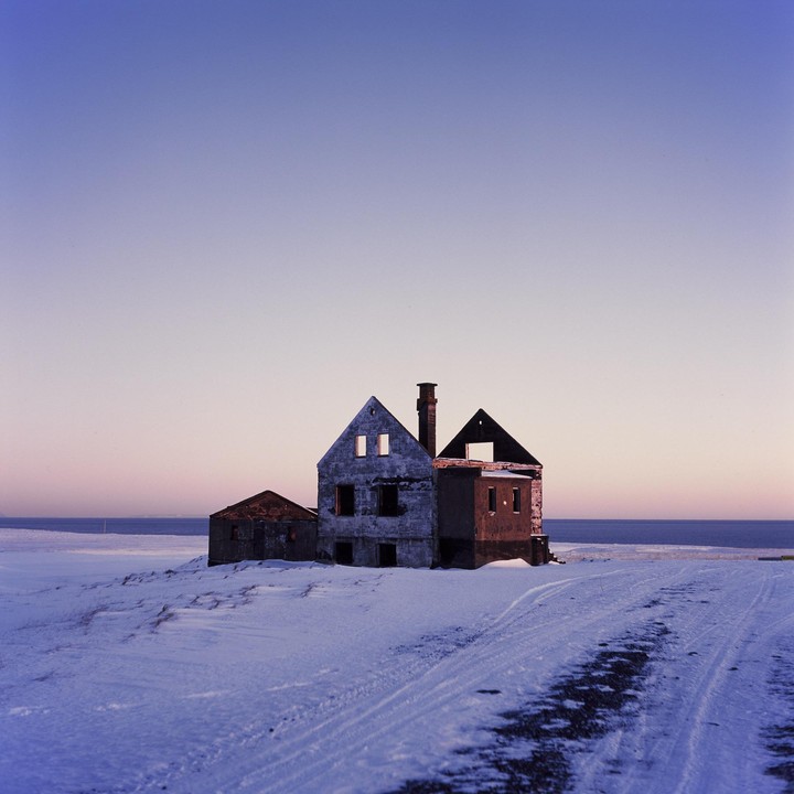 Three abandoned buildings stand on a snow-covered landscape next to the ocean under a clear sky.