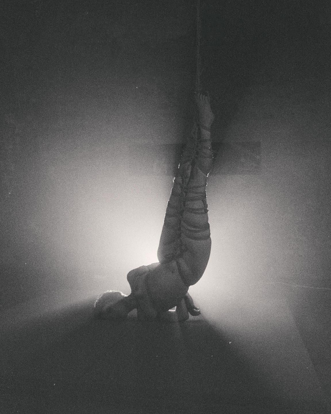 A woman is suspended upside down by ropes in a dark room with a bright light behind her.