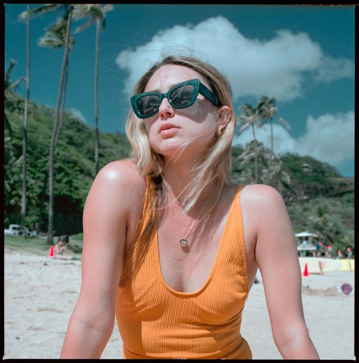 A woman in a yellow swimsuit and sunglasses sits on a sandy beach with palm trees and a cloudy sky in the background.