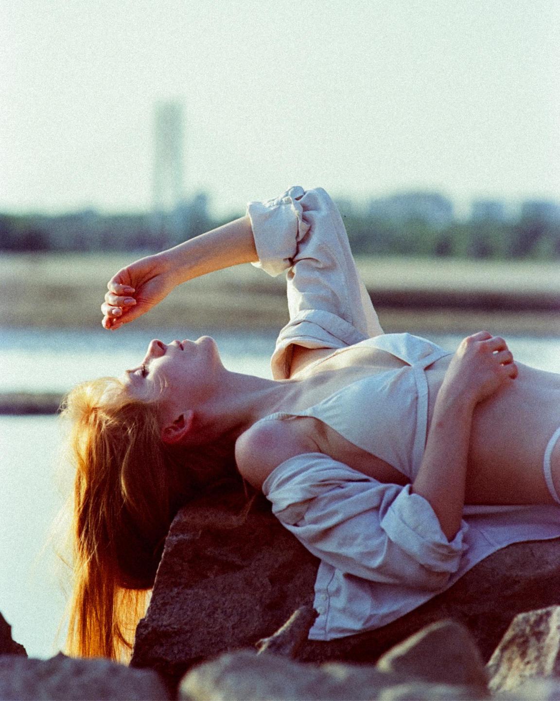 A woman with long red hair lies on rocks by the water, looking up with one arm raised.