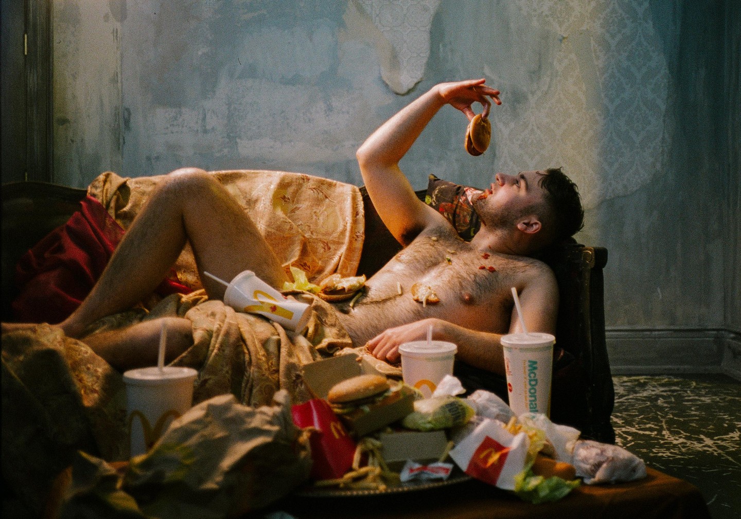 A man reclines on a couch, holding a burger bun above his mouth, surrounded by fast food.