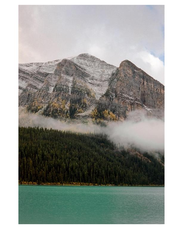 A turquoise lake sits in front of a dense forest and a large, snow-dusted mountain with clouds.