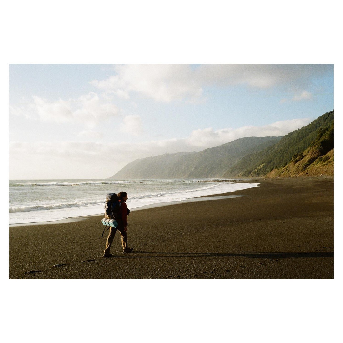 A person with a large backpack walks along a dark sand beach next to an ocean with waves, with forested mountains in the background.