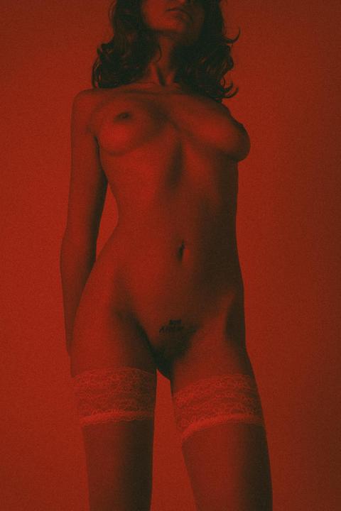 A woman stands nude with her body illuminated by red light, wearing white lace thigh-high stockings.