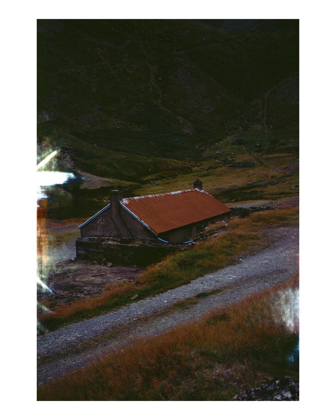 An abandoned mine building with a rusty roof sits beside a gravel road in a mountainous landscape.