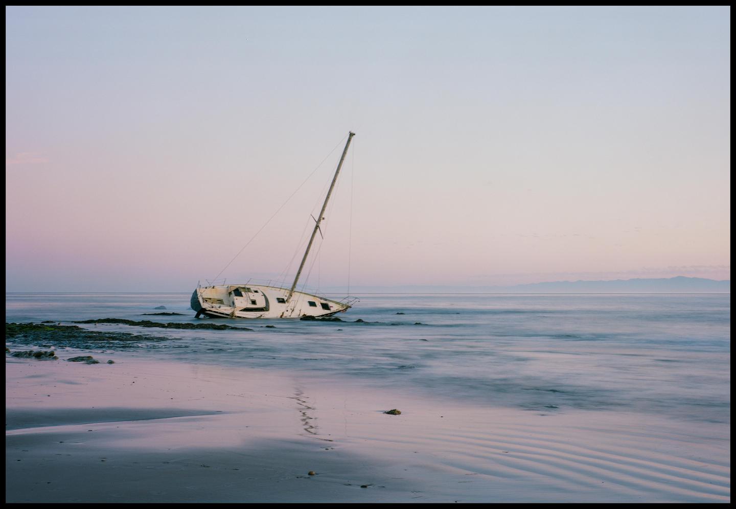 A wrecked sailboat sits on a rocky beach with mountains in the distance under a pastel sky.