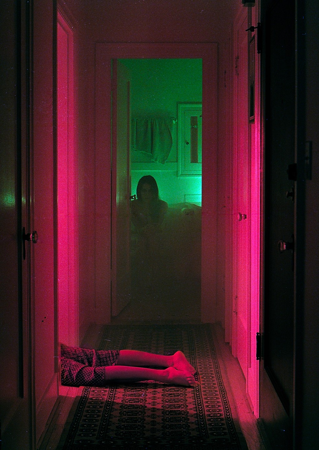 A person's legs are visible on a patterned rug in a hallway lit with pink light, while a figure sits in a green-lit room at the end of the hallway.