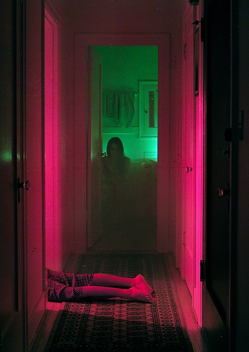 A person's legs are visible on a patterned rug in a hallway lit with pink light, while a figure sits in a green-lit room at the end of the hallway.