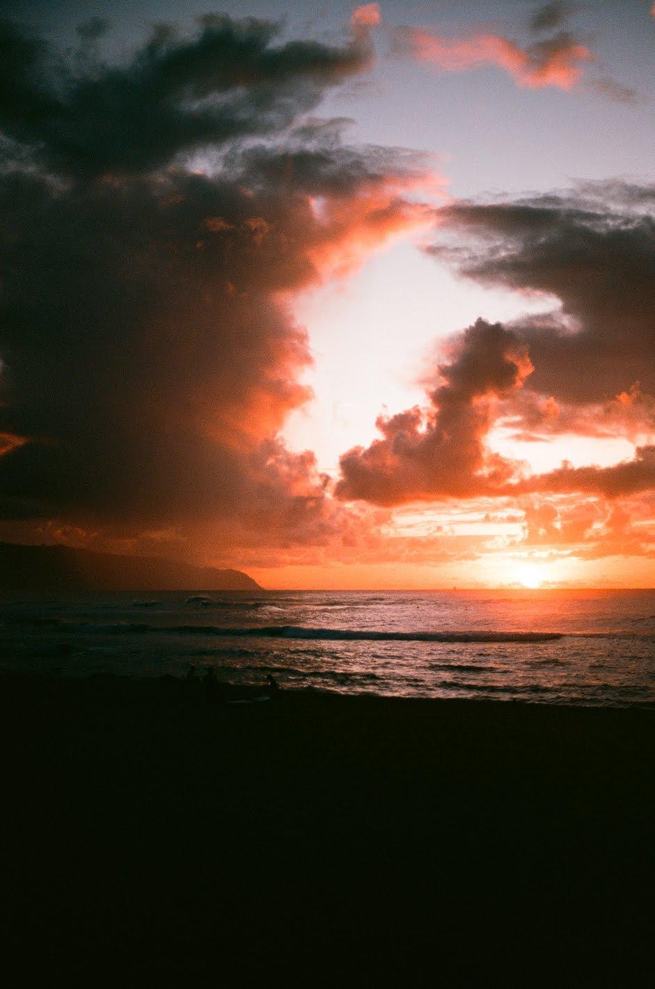 A vibrant sunset over the ocean with dark clouds and a silhouetted beach in the foreground.