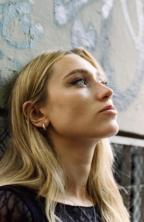 A woman with long blonde hair and blue eyes looks up while leaning against a graffiti-covered wall.
