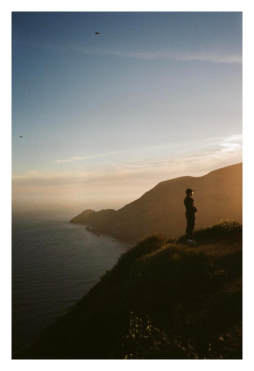 A man stands on a grassy cliff overlooking the ocean and mountains during golden hour.