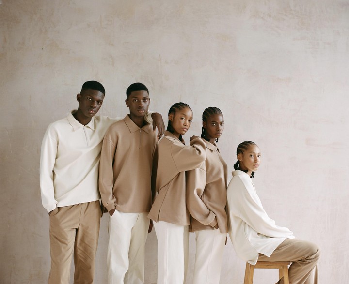 Five people wearing neutral-colored clothing stand and sit in a line against a textured wall.