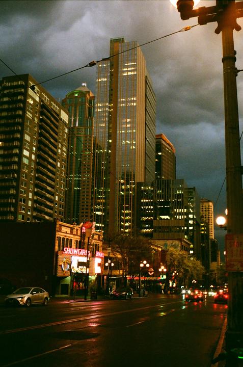 A street scene at night shows tall buildings with illuminated windows under a cloudy sky, with a neon sign for "SHOWGIRLS" visible on a lower building.