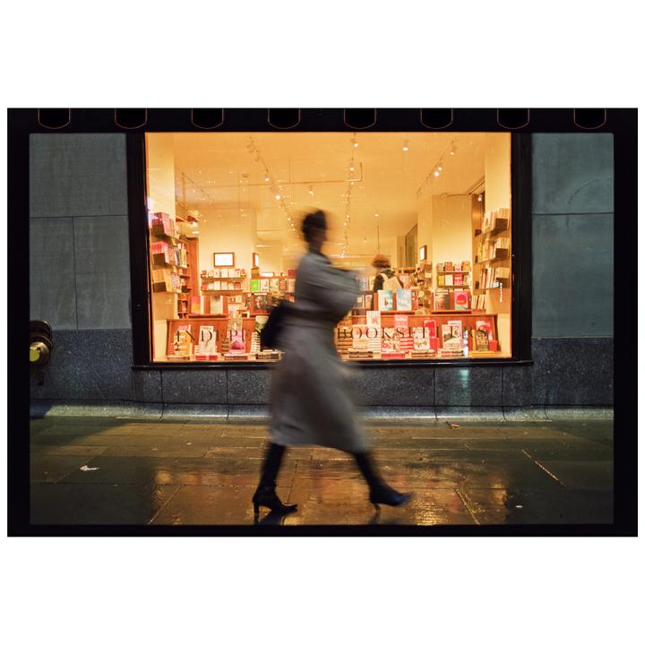 A blurry woman walks past a brightly lit bookstore window at night.