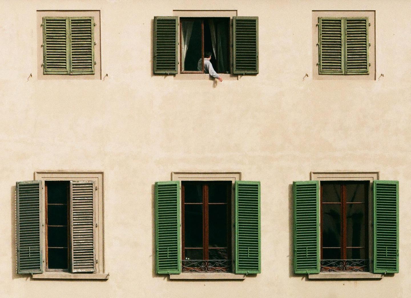 A person leans out of an open window with green shutters on a building facade.