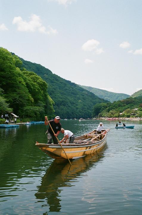 Three men are in a wooden boat on a river surrounded by lush green mountains.