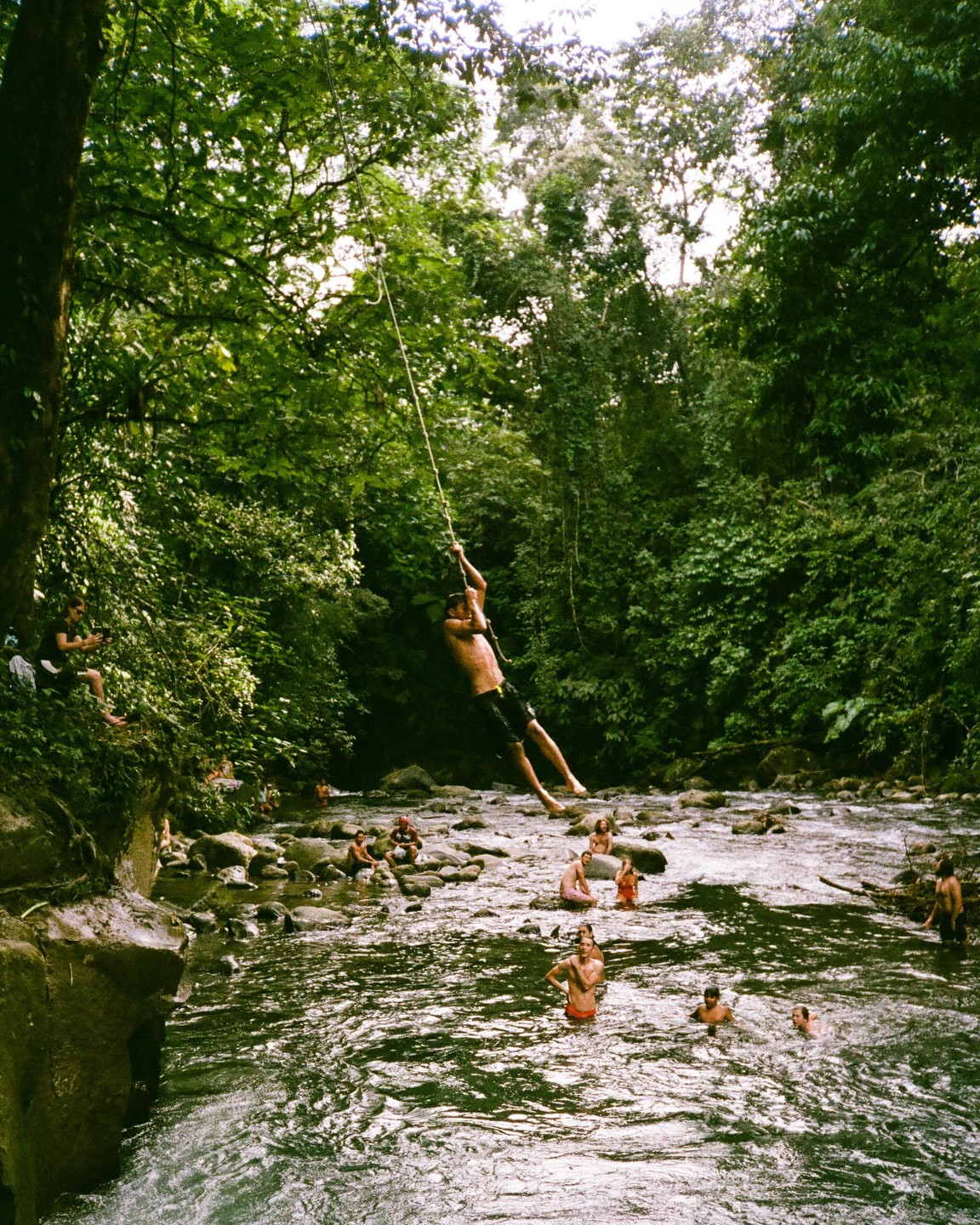 A man swings on a rope over a rocky river with other people swimming and sitting on rocks in a lush, green forest.