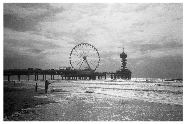 A man and child stand on a beach with a pier and ferris wheel in the background.