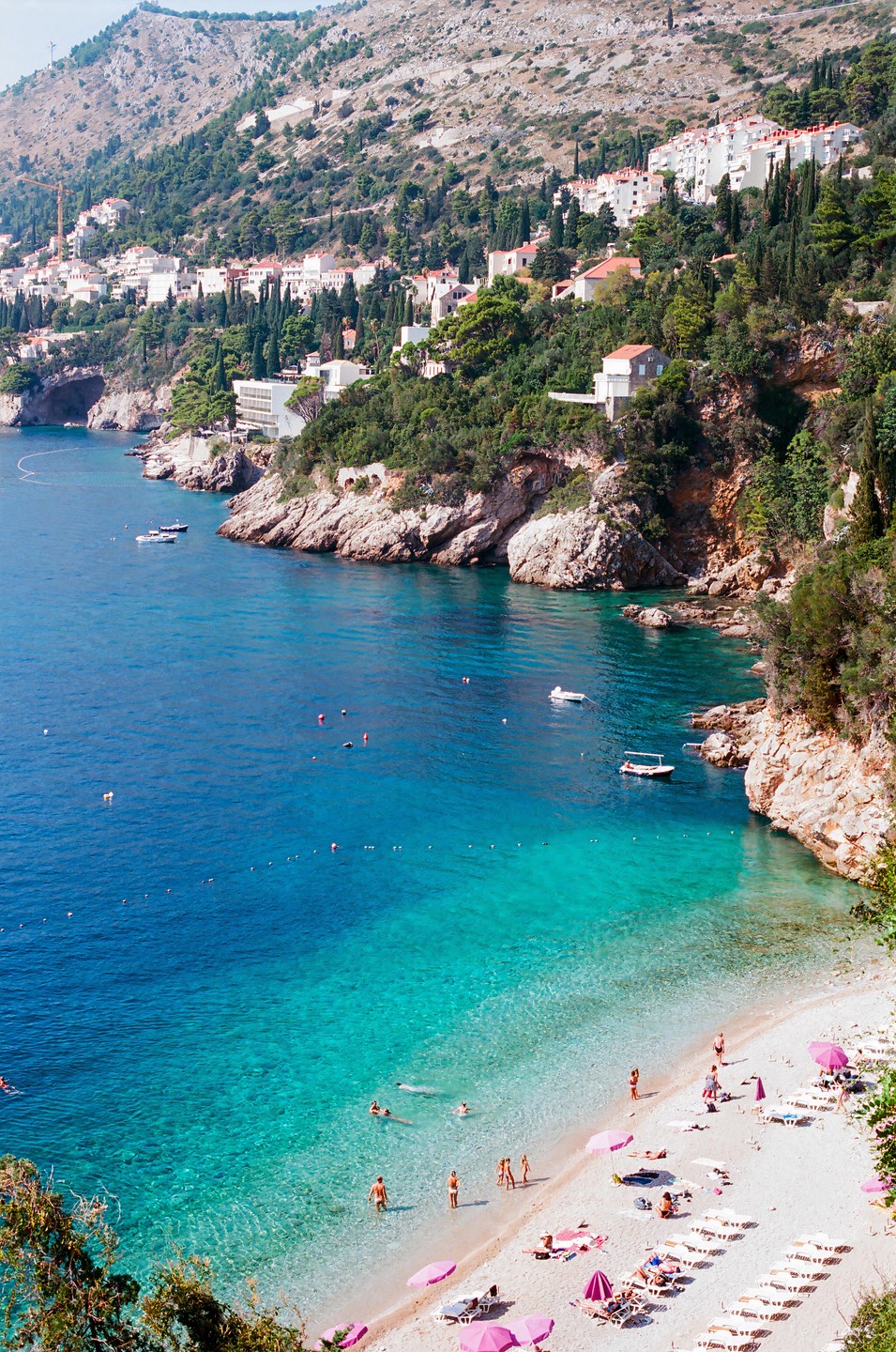 A high-angle view shows a beach with pink umbrellas and people swimming in turquoise water next to a rocky coastline with houses on a hill.