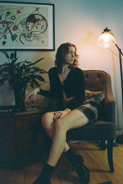 A woman sits in a chair next to a plant and a lamp, looking away from the camera.