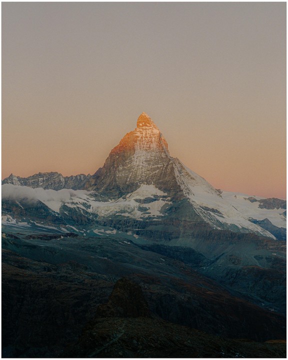 The Matterhorn mountain peak is illuminated by the first light of dawn, with snow-covered slopes and a vast rocky landscape below.