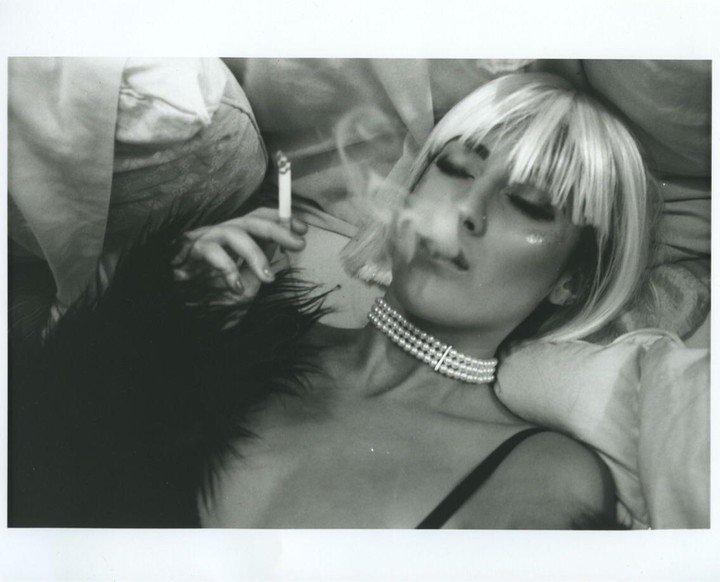 A woman with a pearl necklace and a blonde wig lies down and smokes a cigarette.