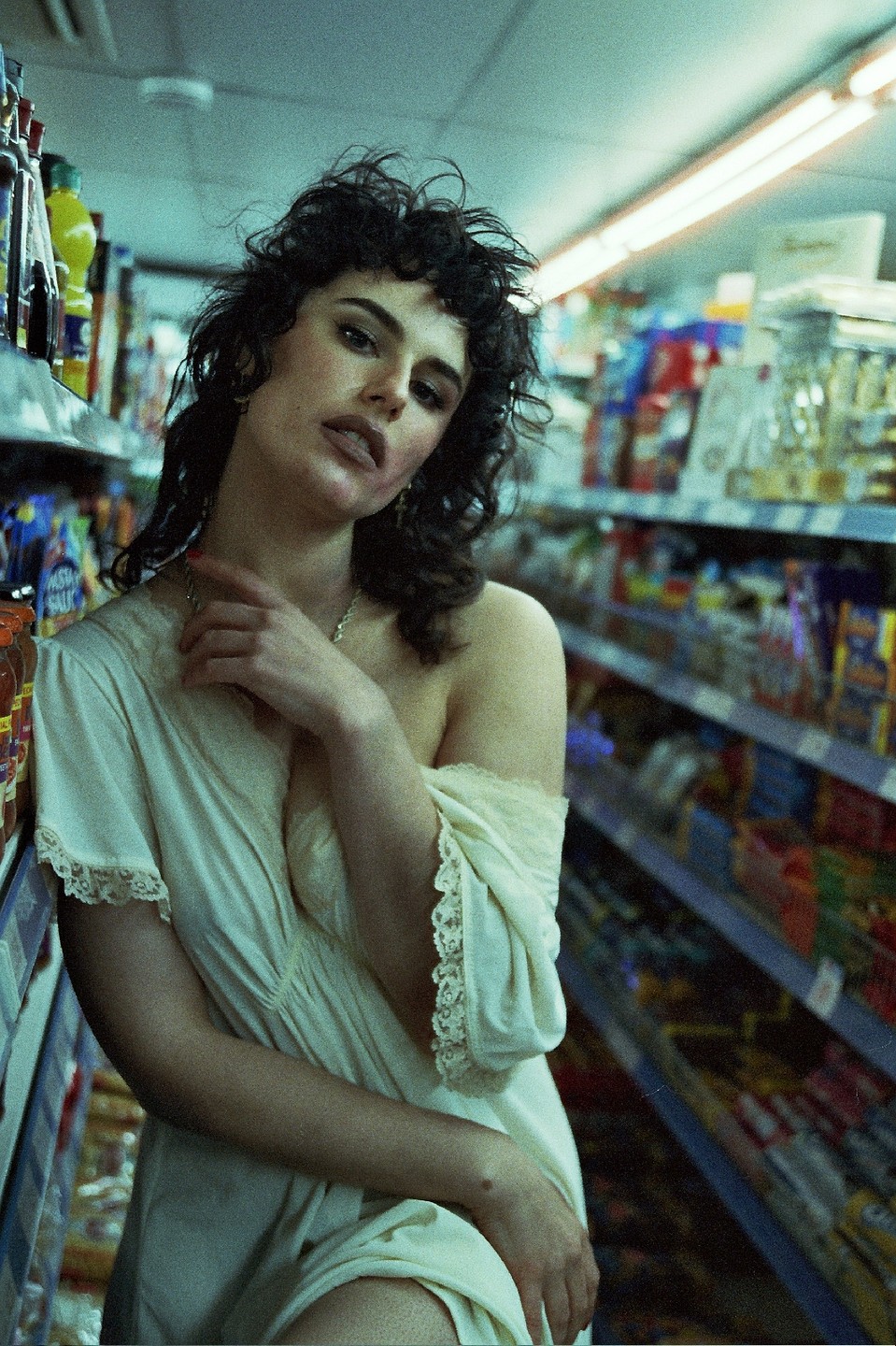 A woman with messy hair and a slip dress poses in a grocery store aisle.