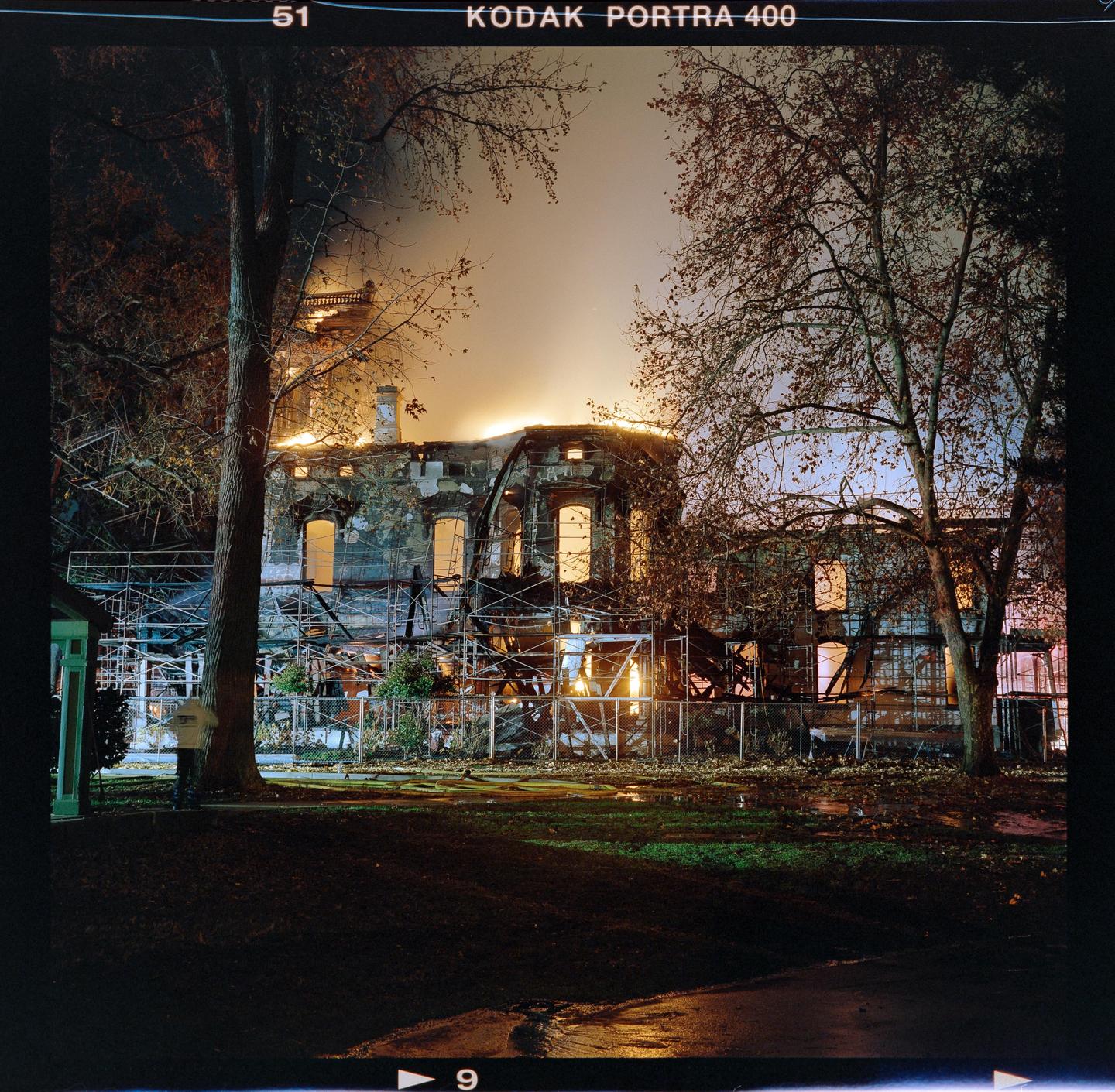 A large mansion is engulfed in flames at night, with scaffolding visible around its exterior.