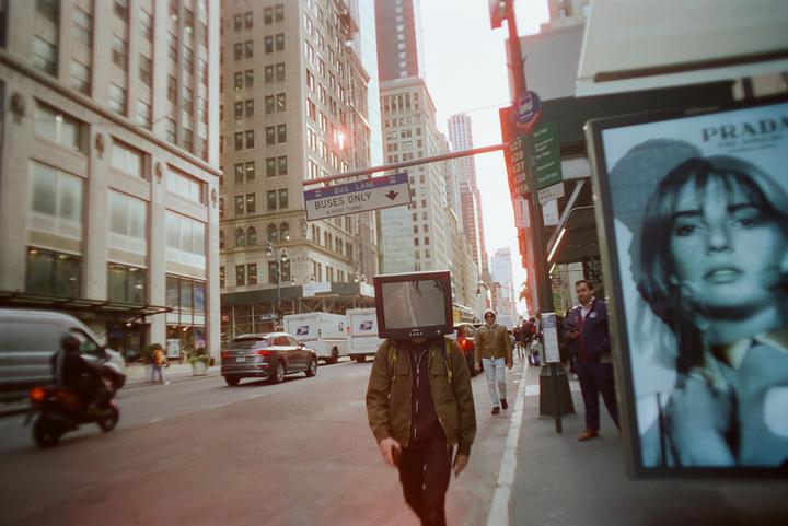 A person with a television for a head walks down a city street past a Prada advertisement.
