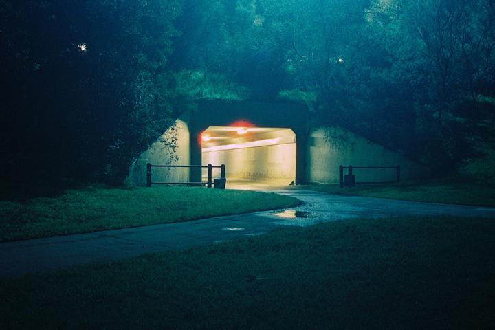 A path leads to a brightly lit tunnel entrance surrounded by dark foliage at night.